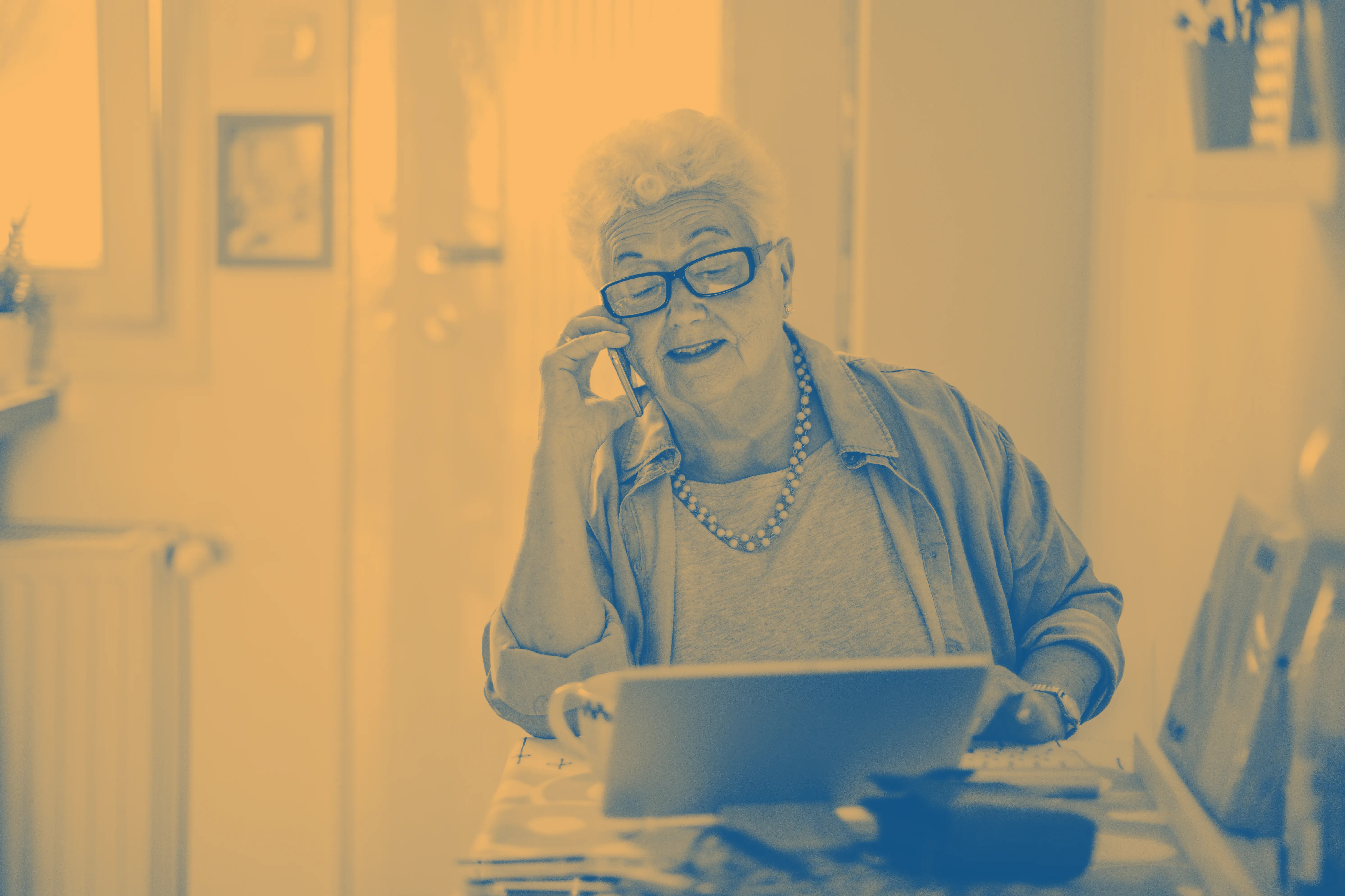 Phot of a mature lady sat at a table speaking on the phone while using a laptop. The lady is smiling